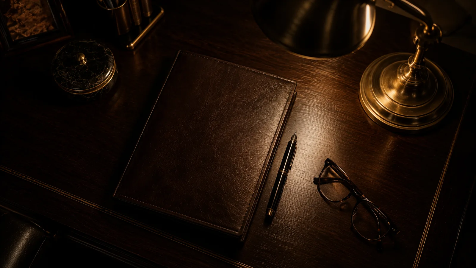 A dark wooden desk with a brass lamp, reading glasses, and a closed leather folder in warm light