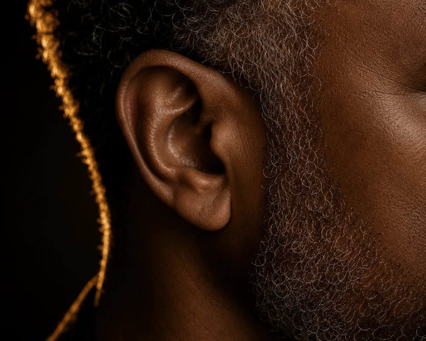 Close detail of a man's ear, jaw, and silver beard, rim-lit in gold